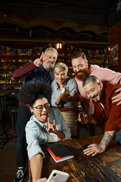 african american woman showing rock sign while taking selfie with multiethnic workmates in pub - Photo, Image