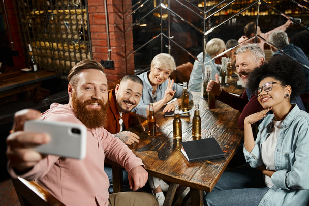 felice uomo barbuto scattare foto con compagni di lavoro multietnici vicino bottiglie di birra sul tavolo di legno nel bar - Foto, immagini
