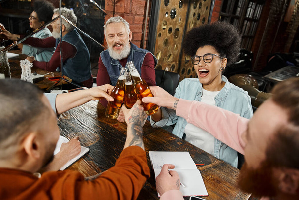 compagni di lavoro multietnici felicissimi che si accarezzano bottiglie di birra mentre riposano nel pub dopo il lavoro, ora di festa - Foto, immagini