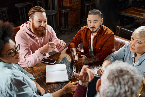 multi-etnische collega 's zitten in pub en bespreken business project in de buurt van bierflessen en notebook - Foto, afbeelding