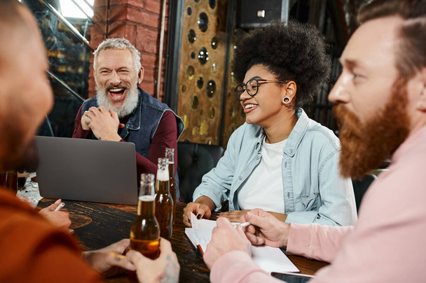 hombre barbudo emocionado riendo cerca de la computadora portátil y compañeros de trabajo multiétnicos con botellas de cerveza hablando en el pub - Foto, Imagen