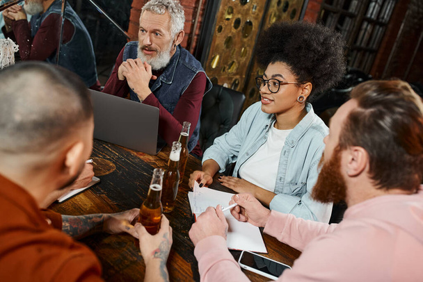 colegas de negocios multiétnicos sentados en un pub cerca de la computadora portátil mientras planifican un nuevo proyecto de inicio - Foto, Imagen