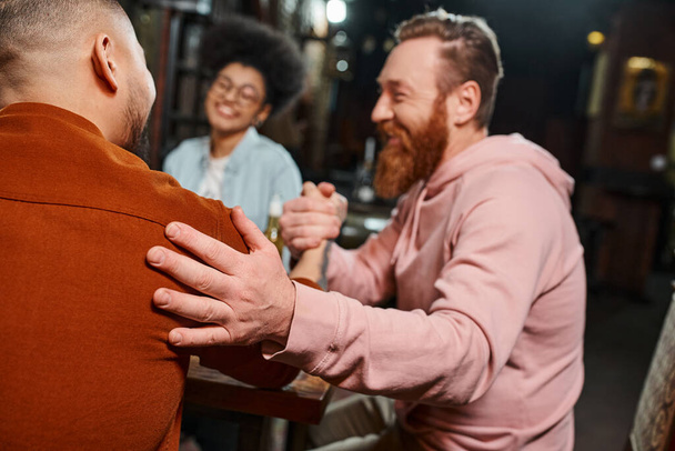 homem barbudo satisfeito apertando as mãos com colega no pub perto de mulher americana africana borrada - Foto, Imagem