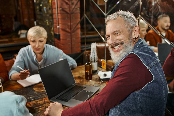 homem barbudo alegre olhando para a câmera perto de laptop e colegas multiétnicos planejamento projeto no pub - Foto, Imagem
