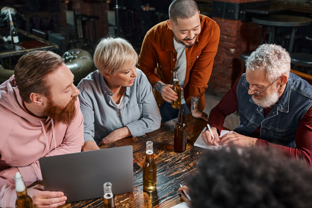 une équipe multiethnique diversifiée regardant un collègue écrire dans un carnet pendant une réunion dans un pub après le travail - Photo, image