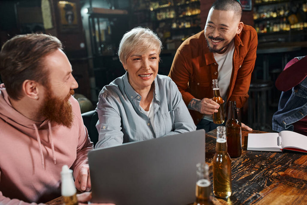 feliz mujer de mediana edad sonriendo cerca de colegas barbudos multiétnicos proyecto de planificación en el pub - Foto, Imagen