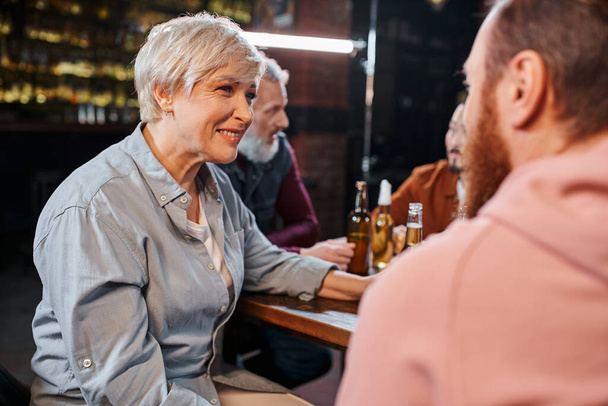 feliz mulher de meia-idade ouvindo colega barbudo perto turvo equipe multiétnica falando no pub - Foto, Imagem