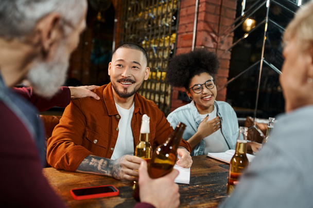 colegas de negócios multiétnicos alegres segurando garrafas de cerveja e falando sobre novo projeto no pub - Foto, Imagem