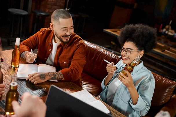 cheerful african american woman with beer bottle near asian colleague sitting with notebook in pub - Photo, Image
