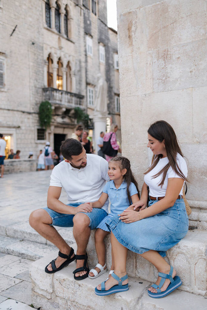 Young family of dad mom and daughter sits on stone in old town. Beautiful people. Denim style. High quality photo - Photo, Image
