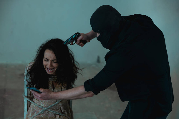 Crimineel in zwarte kleren bedreigt de dame met een pistool en dwingt haar te spreken op de mobiele telefoon. Misdaad, ontvoering, geweld - Foto, afbeelding