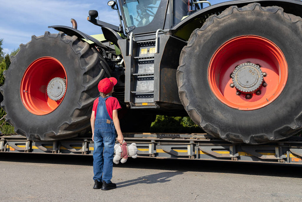 Kind begutachtet einen riesigen Traktor mit großen Rädern, der zum Transport auf einen Lastwagen geladen wird. Unerkennbarer Junge in Jeans-Overalls steht mit dem Rücken im Rahmen. - Foto, Bild