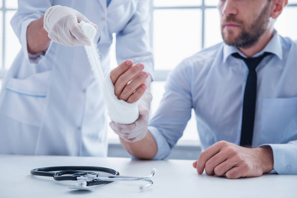 geschnittenes Bild einer Ärztin, die dem Mann bei der Arbeit in ihrem Büro die verletzte Hand bandagiert - Foto, Bild