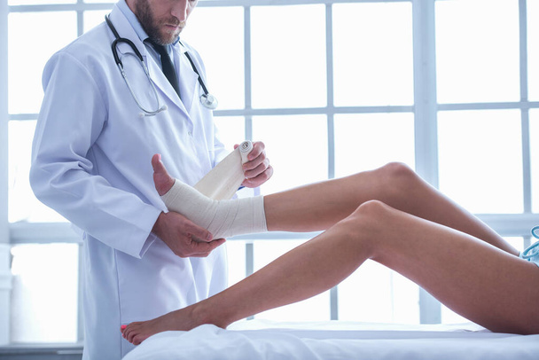 geschnittenes Bild eines gut aussehenden Arztes, der das verletzte Bein einer Frau bei der Arbeit in seinem Büro bandagiert - Foto, Bild
