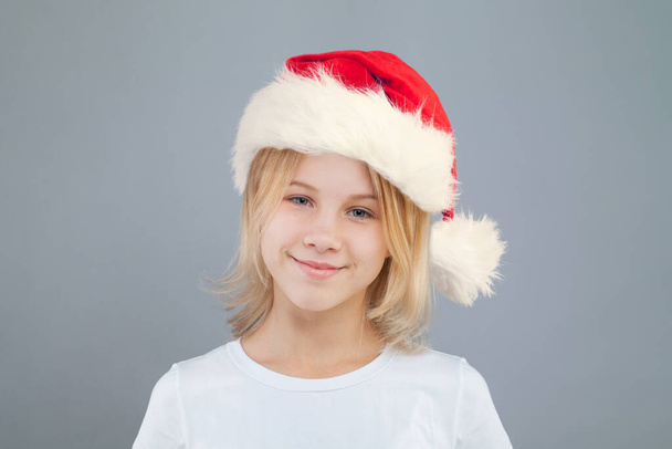 Weihnachtsmädchen mit süßem Lächeln trägt Weihnachtsmütze auf grauem Hintergrund - Foto, Bild