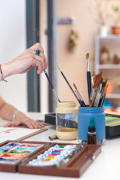 Die Hand der Illustratorin taucht ihren Pinsel in einen Topf mit Wasser, um ihre Aquarellarbeiten zu verrichten.  - Foto, Bild