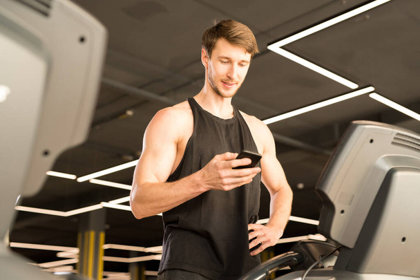 Young man in sportswear communicating online using his mobile phone while standing on treadmill - Foto, afbeelding