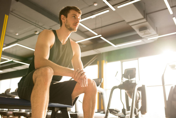 Young athlete in tank top and shorts sitting in gym and resting after sports training - 写真・画像