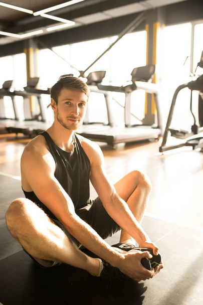 Νεαρός μυώδης άνδρας με αθλητικά ρούχα κάθεται στο πάτωμα στο στρώμα άσκησης και κοιτάζοντας την κάμερα κατά την άσκηση στο γυμναστήριο - Φωτογραφία, εικόνα