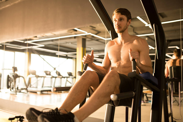 Muscular shirtless man doing exercises on fitness machine in gym - Foto, afbeelding