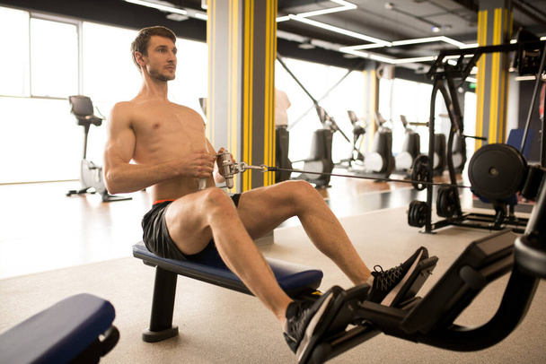 Shirtless muscular athlete sitting and pulling the weight on himself while training on rowing machine in gym - Φωτογραφία, εικόνα