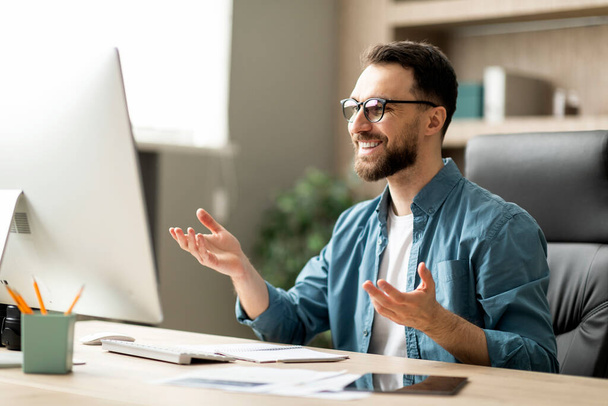 Hombre atractivo empresario haciendo videollamada mientras está sentado en el escritorio en la oficina, hombre de negocios milenario mirando el monitor de la computadora y sonriendo, disfrutando de la comunicación en línea, primer plano - Foto, Imagen