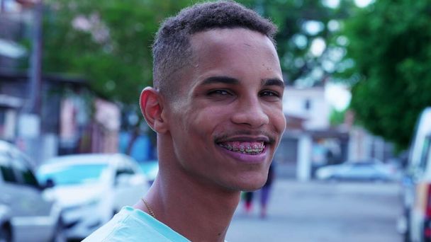一人の幸せな自信を持ったブラジルの若い男性は,バックグラウンドで都市のストリートと立っている間,カメラで微笑みます, クローズアップ顔の肖像画 - 写真・画像