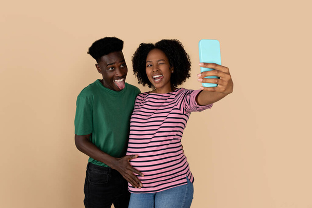 Usmívající se tisíciletá černá rodina těšit těhotenství, vzít selfie na smartphone, mrkne a vyčnívá jazyk, izolované na béžové pozadí, studio. Životní styl, zábava a očekávání miminko, foto - Fotografie, Obrázek