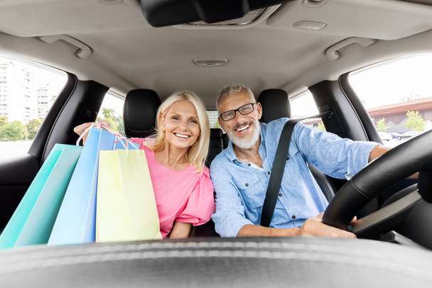 Weseli, piękni starsi małżonkowie jadący do domu po zakupach w centrum handlowym, uśmiechnięci. Szczęśliwy starszy mężczyzna i kobieta siedzący w samochodzie, trzymający kolorowe zakupy torb na zakupy. Czarny piątek, wyprzedaż sezonowa - Zdjęcie, obraz