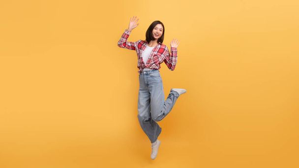 Iloinen nuori aasialainen nainen hyppää ilmassa ja hymyilee kameralle, innoissaan nainen rento asu hauskaa keltainen studio tausta, täyspitkä, kopioi tilaa - Valokuva, kuva