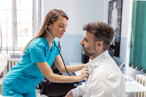Pečující běloška lékař pomocí fonendoskopu vyšetřuje mužskou srdeční frekvenci pacienta po konzultaci v nemocnici. Žena zdravotní sestra nebo GP používat stetoskop poslouchat mužský srdeční tep na klinice. - Fotografie, Obrázek