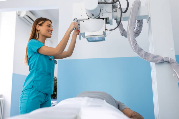 Γιατρός που κοιτάζει το μηχάνημα ακτίνων Χ στην κλινική. Γυναίκα γιατρός στήνει το μηχάνημα για ακτινογραφία πάνω από τον ασθενή. Ακτινολόγος και ασθενής σε αίθουσα ακτινογραφιών. - Φωτογραφία, εικόνα