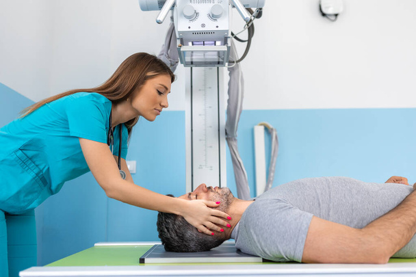 Νεαρή γυναίκα με αυτοπεποίθηση ακτινολόγος γιατρός, που κάνει ακτινογραφία σε άντρα ασθενή ξαπλωμένο στο τραπέζι του μηχανήματος. Γιατρός στέκεται κοντά στον ασθενή κατά τη διάρκεια της διαδικασίας ακτινογραφία κεφαλής - Φωτογραφία, εικόνα