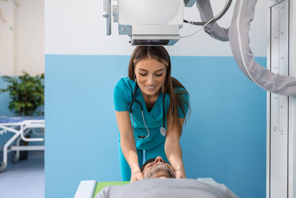 Paciente masculino acostado en la cama mientras enfermera ajustando la moderna máquina de rayos X para escanear su pecho en busca de lesiones y fracturas - Foto, Imagen