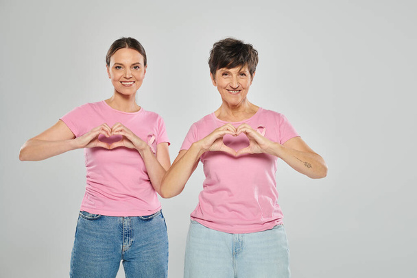 Концепция рака молочной железы, счастливые женщины, смотрящие в камеру и показывающие сердце руками на сером фоне - Фото, изображение