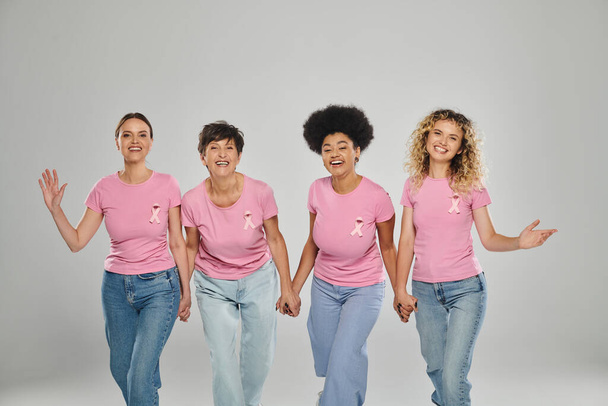 breast cancer awareness, joyful multicultural women with pink ribbons walking together on grey - Фото, изображение