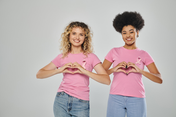 breast cancer awareness, happy multicultural women with pink ribbons on grey backdrop, heart sign - Photo, Image