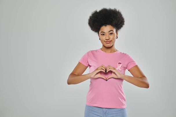 Göğüs kanseri farkındalığı, gri arka planda pembe kurdeleli Afro-Amerikan kadın, kalp işareti, gülümseme - Fotoğraf, Görsel