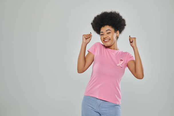 świadomość raka piersi, podekscytowana Afroamerykanka z różową wstążką na szarym, bez raka - Zdjęcie, obraz