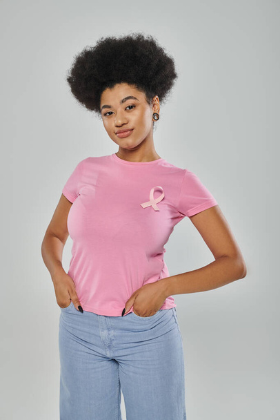 conciencia de cáncer de mama, sonrisa, mujer afroamericana con cinta rosa en gris, manos en bolsillos - Foto, Imagen