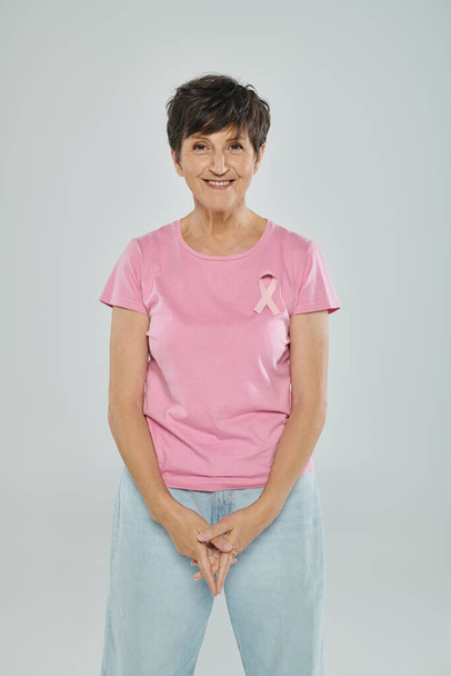 borstkanker bewustzijn, gelukkige vrouw van middelbare leeftijd met roze lint, grijze achtergrond, kankervrij - Foto, afbeelding
