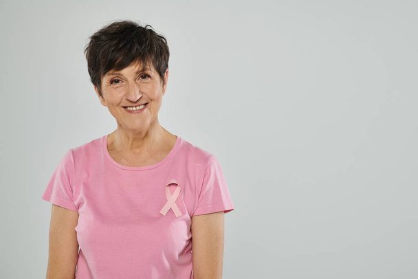 świadomość raka piersi, szczęśliwa kobieta w średnim wieku z różową wstążką, szare tło, portret - Zdjęcie, obraz