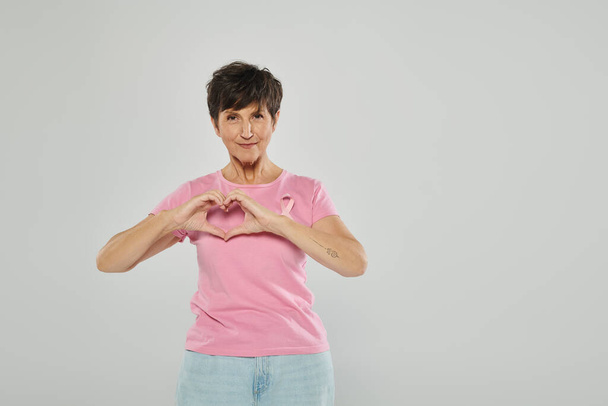 conciencia de cáncer de mama, mujer madura feliz con cinta rosa, fondo gris, retrato, signo del corazón - Foto, imagen
