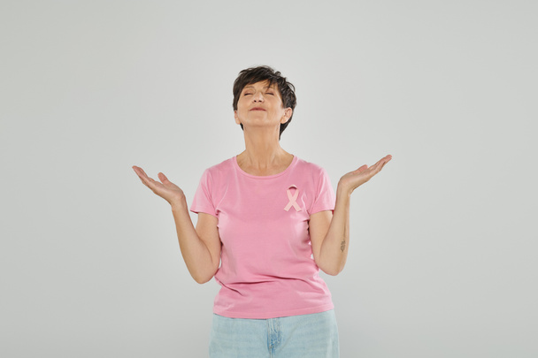 breast cancer awareness, happy mature woman with pink ribbon, grey backdrop, joy, meditation - Photo, Image