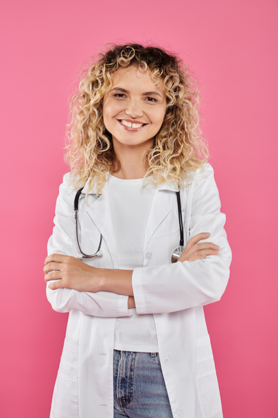 Концепция информированности о раке молочной железы, кудрявая врач смотрит на камеру, розовый фон, улыбка, женщина - Фото, изображение