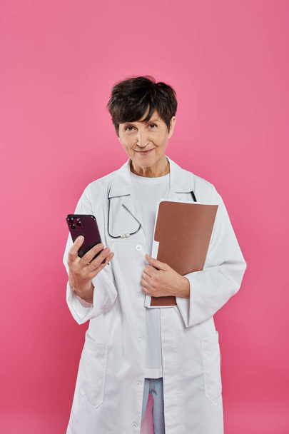 reife Onkologin, Ärztin mit Aktenmappe und Smartphone, Brustkrebsbewusstsein, technisches Alter - Foto, Bild