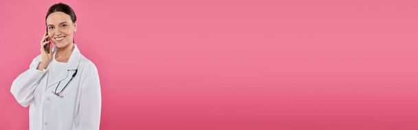 medico sorridente parlando su smartphone mentre in piedi isolato su rosa, banner, concetto di cancro al seno - Foto, immagini