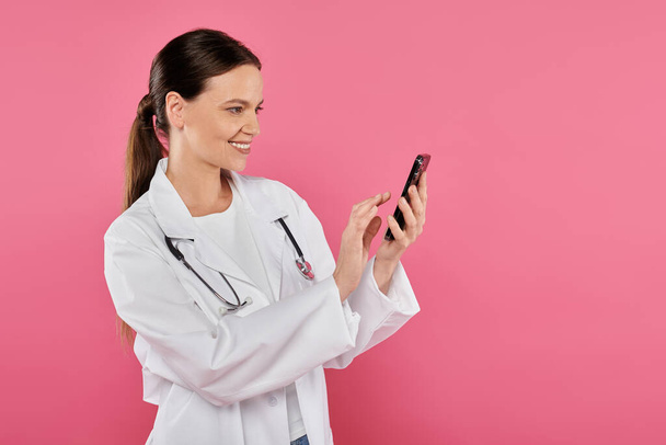 glimlachende vrouwelijke arts in witte jas met behulp van smartphone geïsoleerd op roze, borstkanker concept - Foto, afbeelding