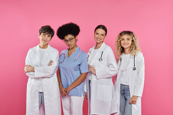 médicos alegres y multiétnicos sonriendo a la cámara aislada en rosa, concepto de cáncer de mama - Foto, Imagen