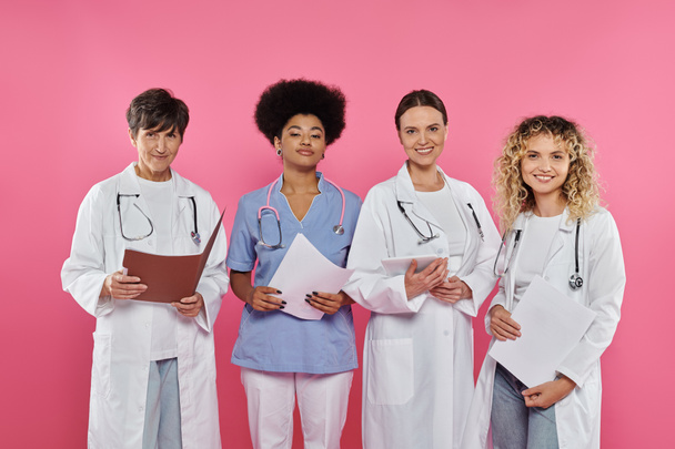 улыбающиеся многонациональные врачи с бумажными папками и цифровыми таблетками, изолированными от розового, рака груди - Фото, изображение